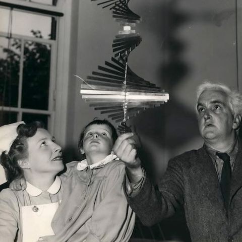 Metal sculpture. Black and white photograph of a senior person holding a metal mobile sculpture and delicately spinning it from its base. A nurse and child gaze up towards the mobile with curious expressions.