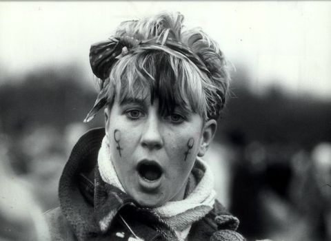 Black and white photograph. Shown from the shoulders up, a person with short hair tied behind a scarf faces us, mouth wide open. Two symbols denoting female are drawn on the cheeks.
