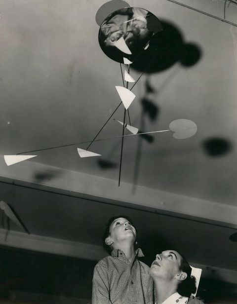Black and white photograph. Nurse holding a child both look up at a mobile sculpture hanging from the ceiling. The sculpture consists of interlocking rods extending down and out in all directions with a few geometric shapes placed throughout the sculpture.